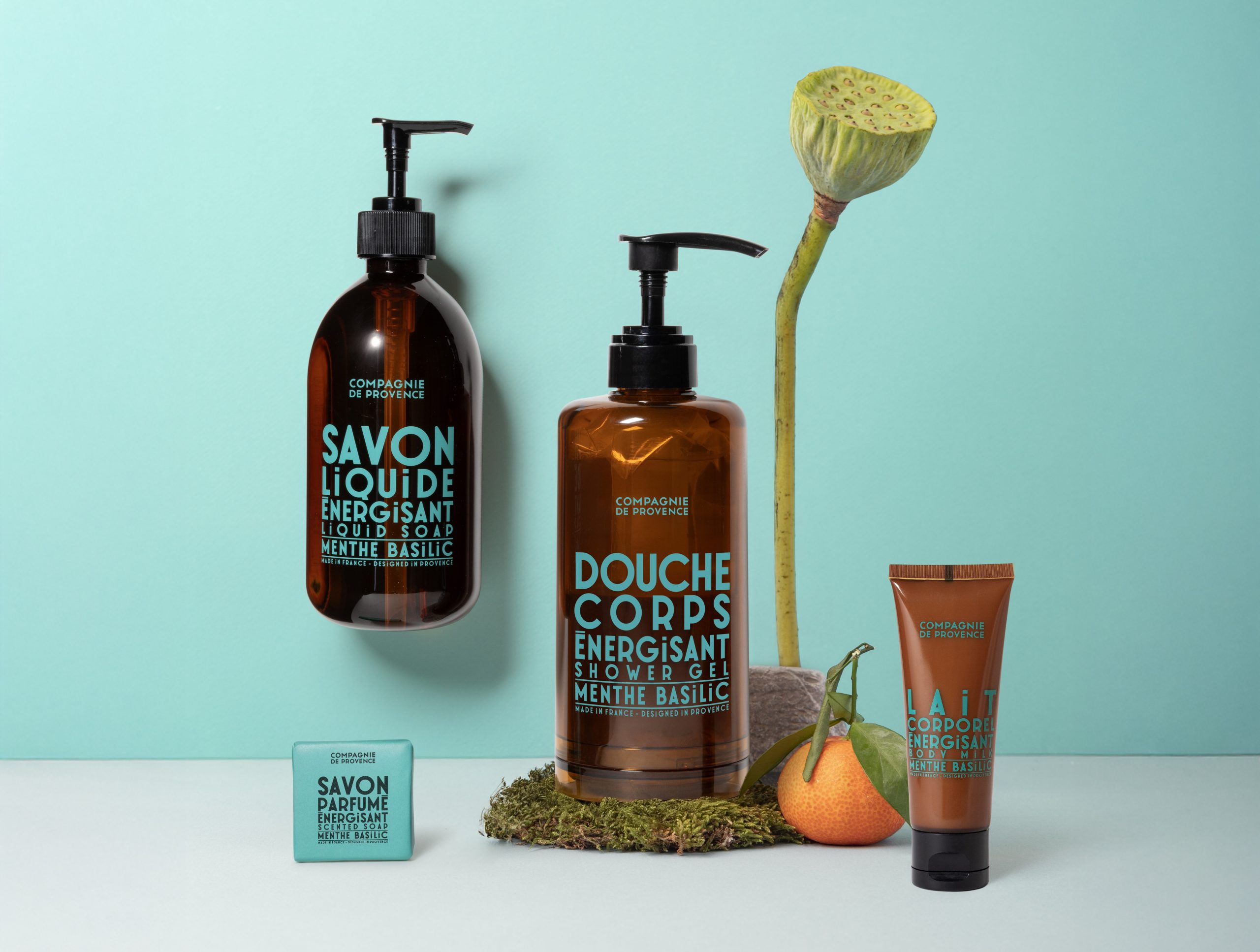 Eine Kollektion belebender Bade- und Körperpflegeprodukte von Compagnie de Provence, darunter Flüssigseife, Duschgel, Körpermilch und duftende Seife, ästhetisch arrangiert mit einer Lotuskapsel, Zitrusfrüchten und Moos vor einem sanften türkisfarbenen Hintergrund.