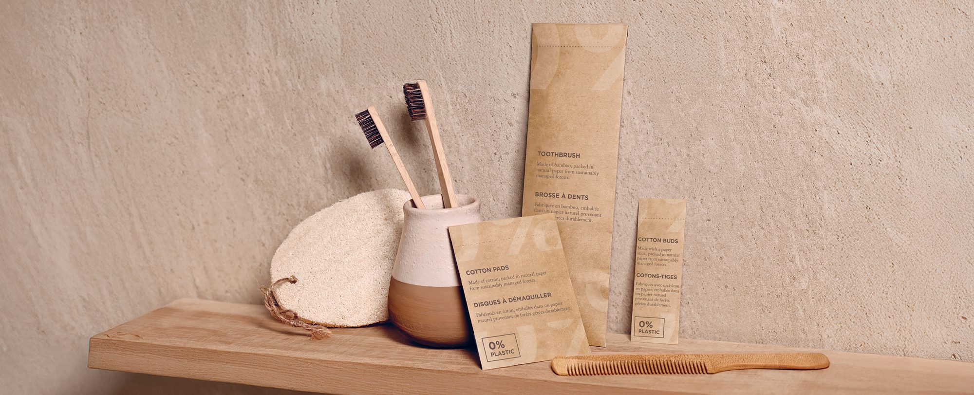 Eco-friendly bathroom products arranged on a wooden shelf, including bamboo toothbrushes, a wooden comb, and biodegradable packaging for cotton swabs and pads. The background features a textured wall for a natural aesthetic.