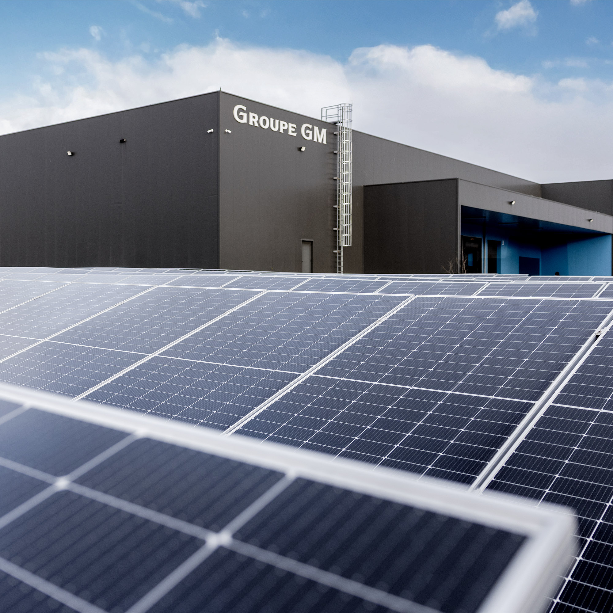 A close-up of solar panels in the foreground with a modern building in the background, featuring the sign "Groupe GM." The sky is partly cloudy.