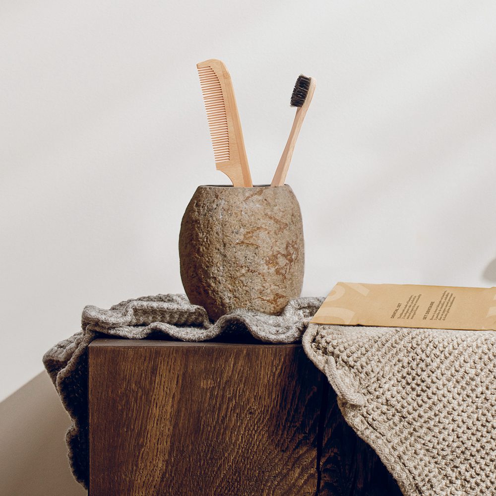 A stone container holds a wooden comb and toothbrush, placed on a textured gray blanket draped over a wooden surface.