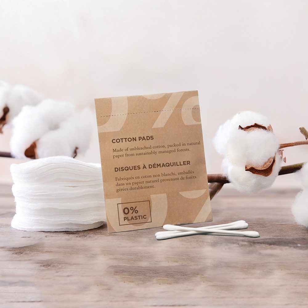 A package of cotton pads made from unbleached cotton is displayed alongside a roll of cotton and bamboo cotton swabs, all resting on a wooden surface. The packaging highlights that it is 100% plastic-free.