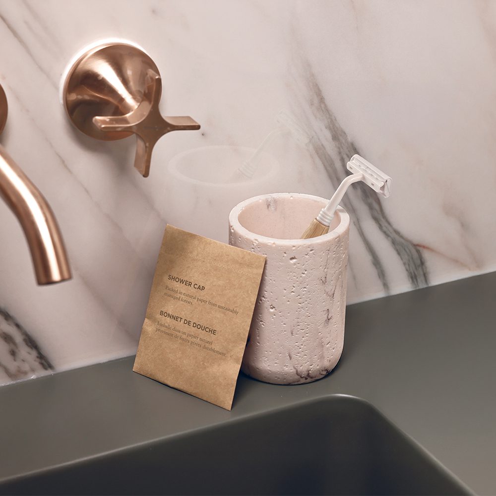 A pink marbled container holds a razor next to a brown paper package labeled "Shower Cap" on a dark green surface, with stylish faucets in the background.