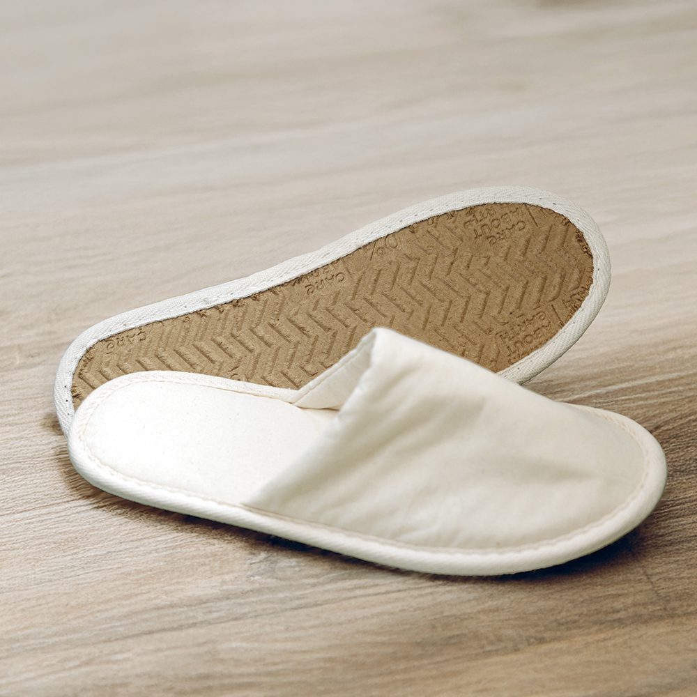A pair of beige fabric slippers with a simple design, resting on a wooden floor. One slipper is upside down, showing its sole.