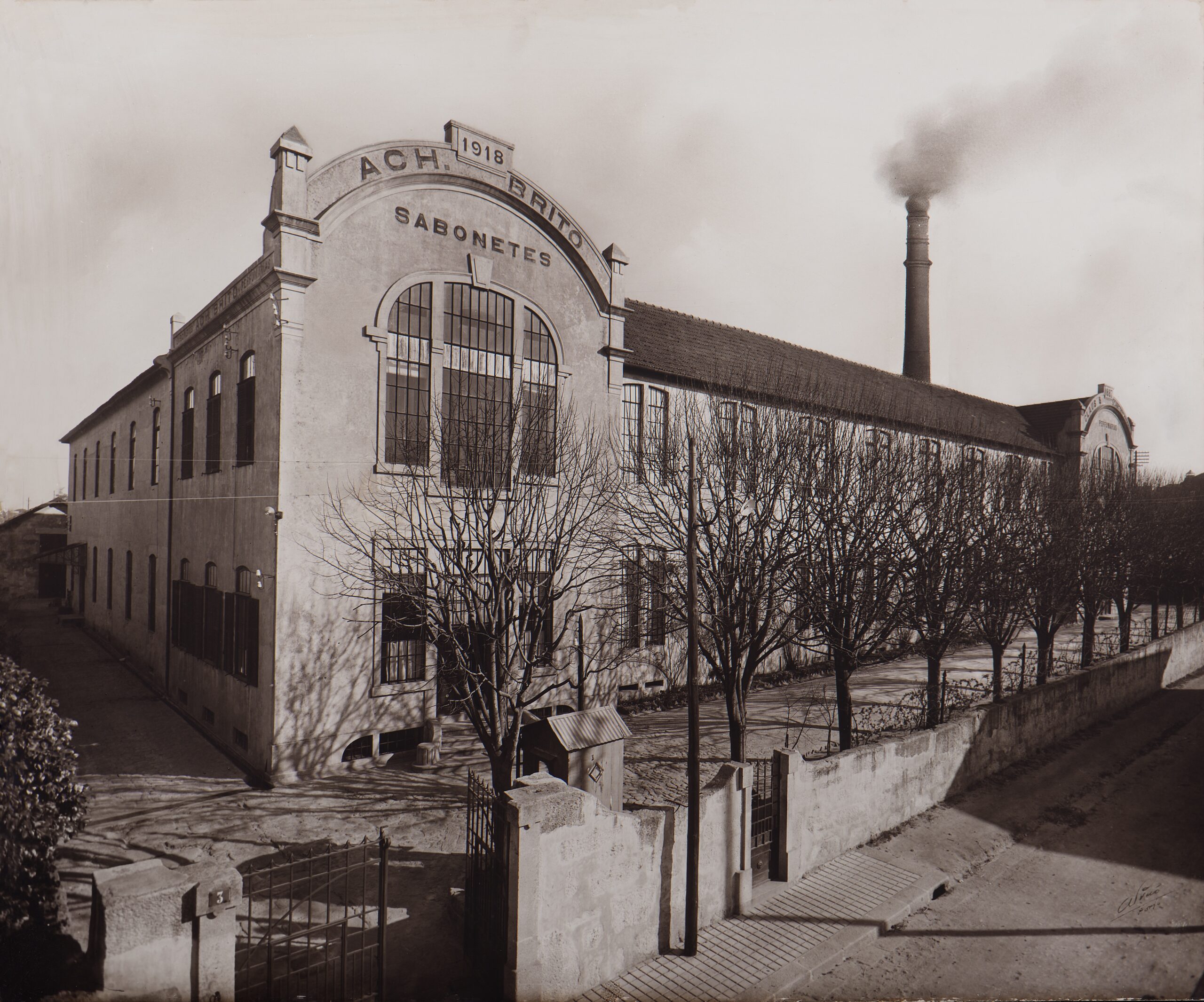 Historic factory building with the year 1918 displayed, featuring a tall chimney emitting smoke and surrounded by bare trees.