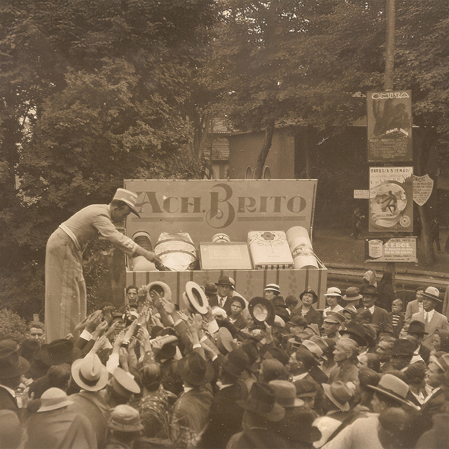 A vintage photo showing a crowd of people reaching out towards a tall vendor handing out products from a large display sign that reads "A. C. H. Brito." The scene is filled with hats and excited faces, suggesting a lively event.