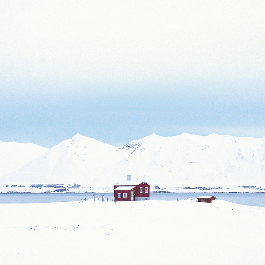 A small red house stands alone on a snowy landscape, surrounded by white mountains and a calm body of water under a pale blue sky.