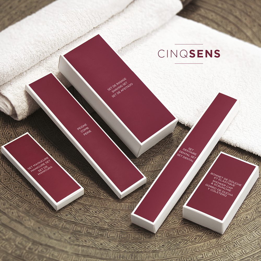 A collection of elegantly packaged spa products in varying sizes, featuring a maroon color scheme, displayed on a textured surface with a white towel in the background. The brand name "CINQ SENS" is prominently featured at the top.