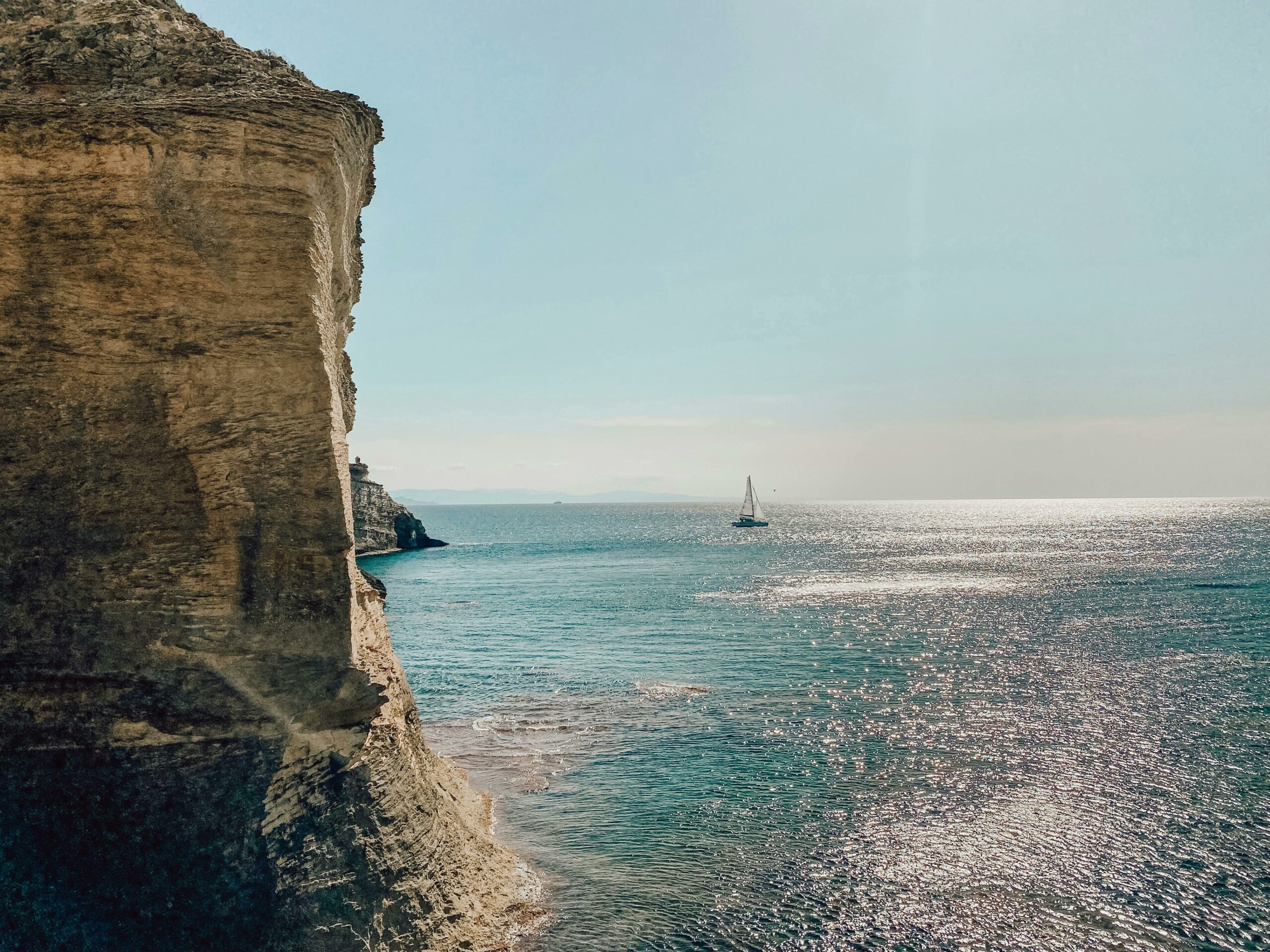 A sailboat glides on shimmering blue waters near a rugged cliff face under a bright sky. The sunlight reflects off the ocean, creating a sparkling effect.