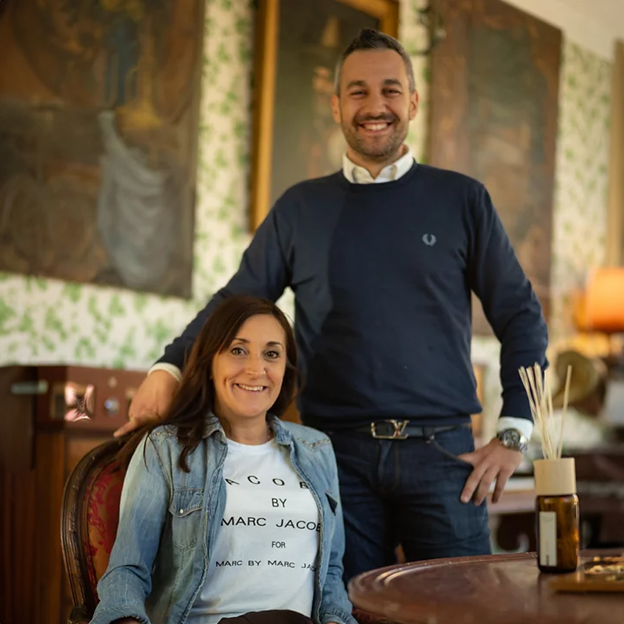 A woman seated in an ornate chair wearing a graphic t-shirt smiles at the camera, while a man stands beside her with a friendly expression, both set against a backdrop of vintage-style decor.