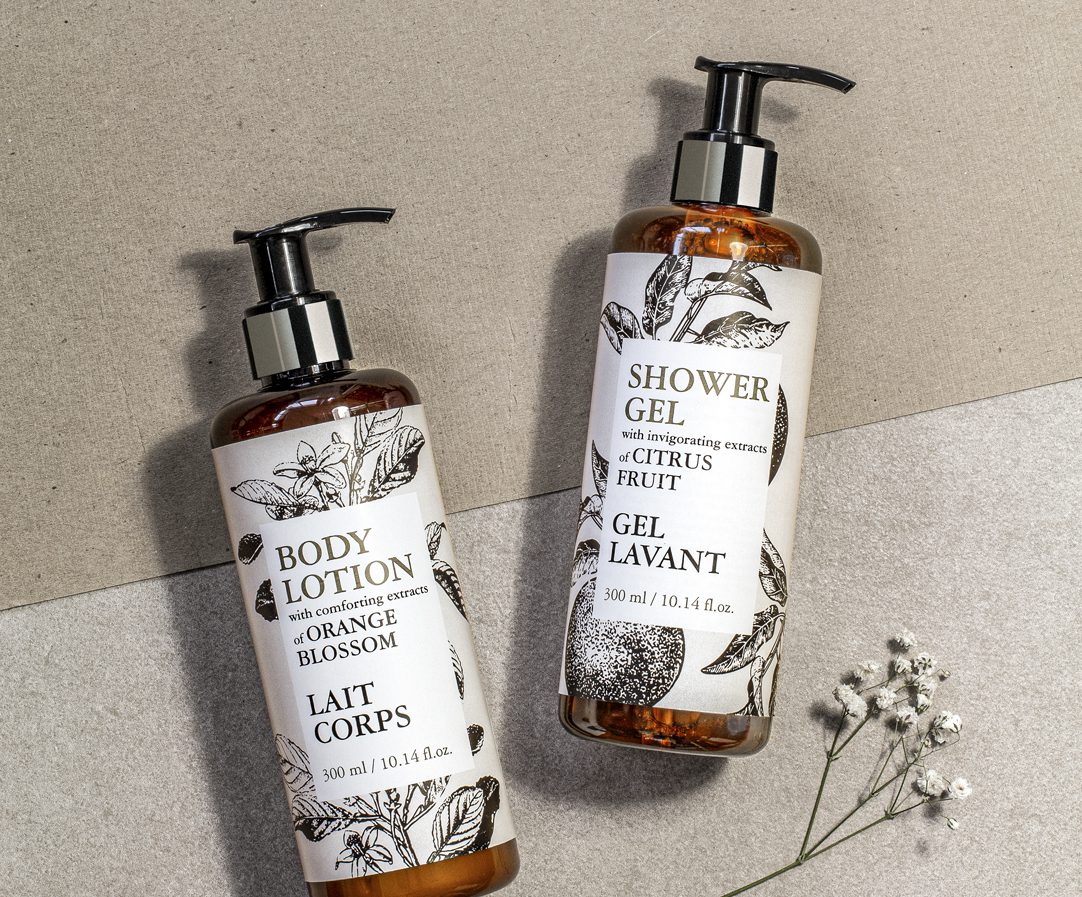 Two bottles of body care products, one labeled "Body Lotion" with orange blossom extract and the other "Shower Gel" with citrus fruit extract, both stylishly packaged with botanical illustrations.