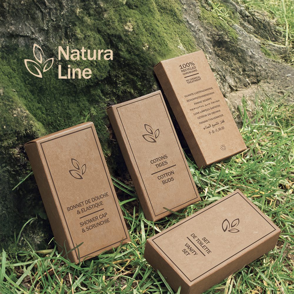 Four eco-friendly product boxes from Natura Line, featuring a shower cap, scrunchie, cotton buds, and a variety set, displayed on grass next to a stone.