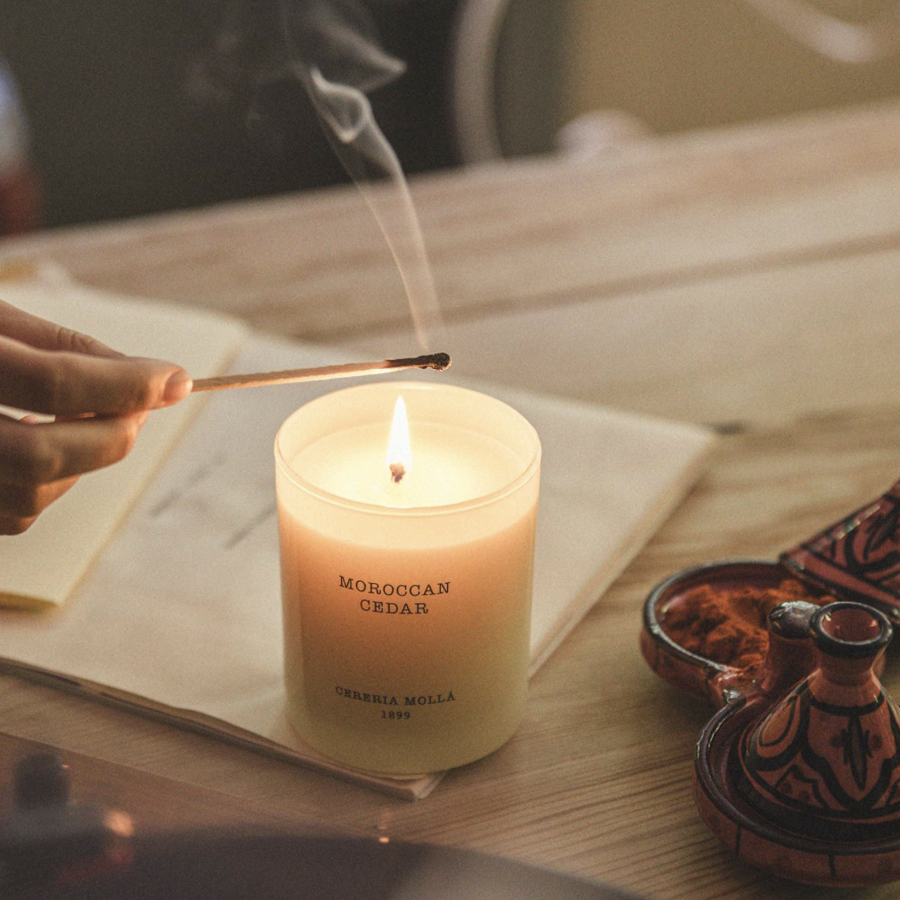 A hand lights a matchstick near a lit candle labeled "Moroccan Cedar," placed on a wooden table alongside an ornate dish. Smoke wisps gently rise from the candle.