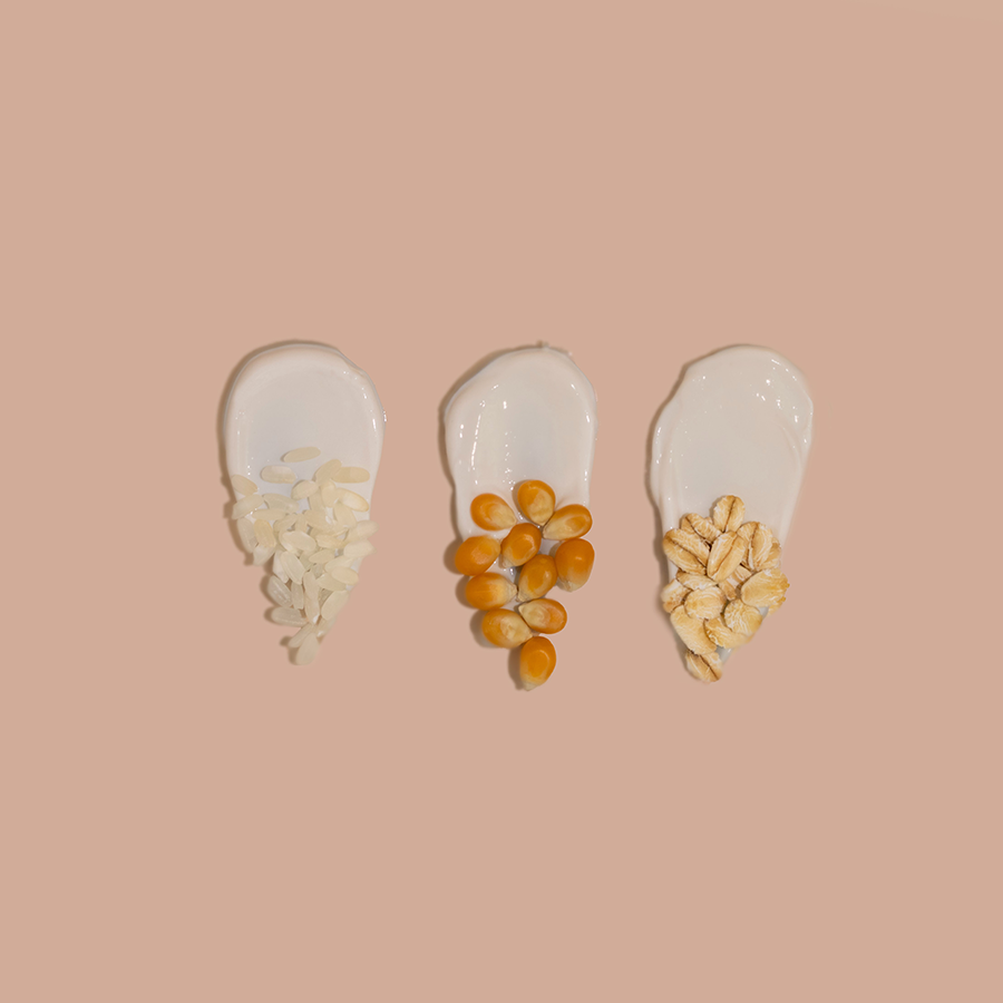 Three spoonfuls of different grains—white rice, orange lentils, and rolled oats—arranged on a soft pink background.