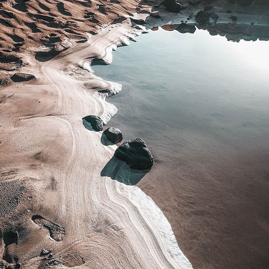 A serene sandbank with smooth textures and gentle waves blends into a calm, reflective pool of water, surrounded by scattered rocks. Sunlight glints off the surface, creating a tranquil ambiance.