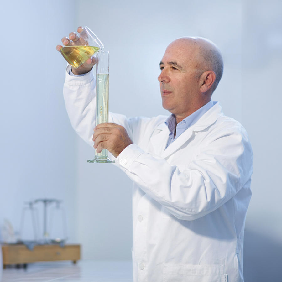 A scientist in a white lab coat pours a yellow liquid from a beaker into a graduated cylinder, focusing intently on the measurement. The laboratory has a bright, sterile appearance.