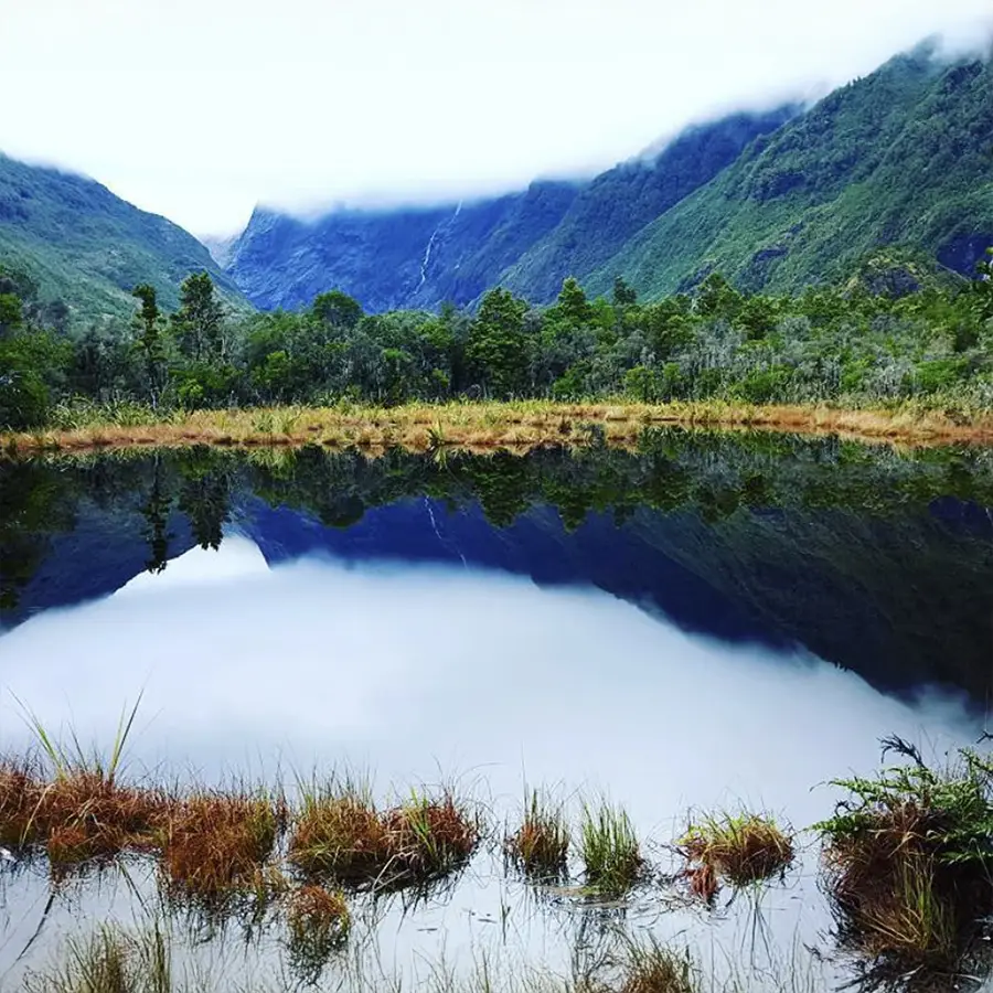 A serene landscape featuring a still lake reflecting the surrounding mountains and lush greenery, with low-lying clouds adding to the misty atmosphere.