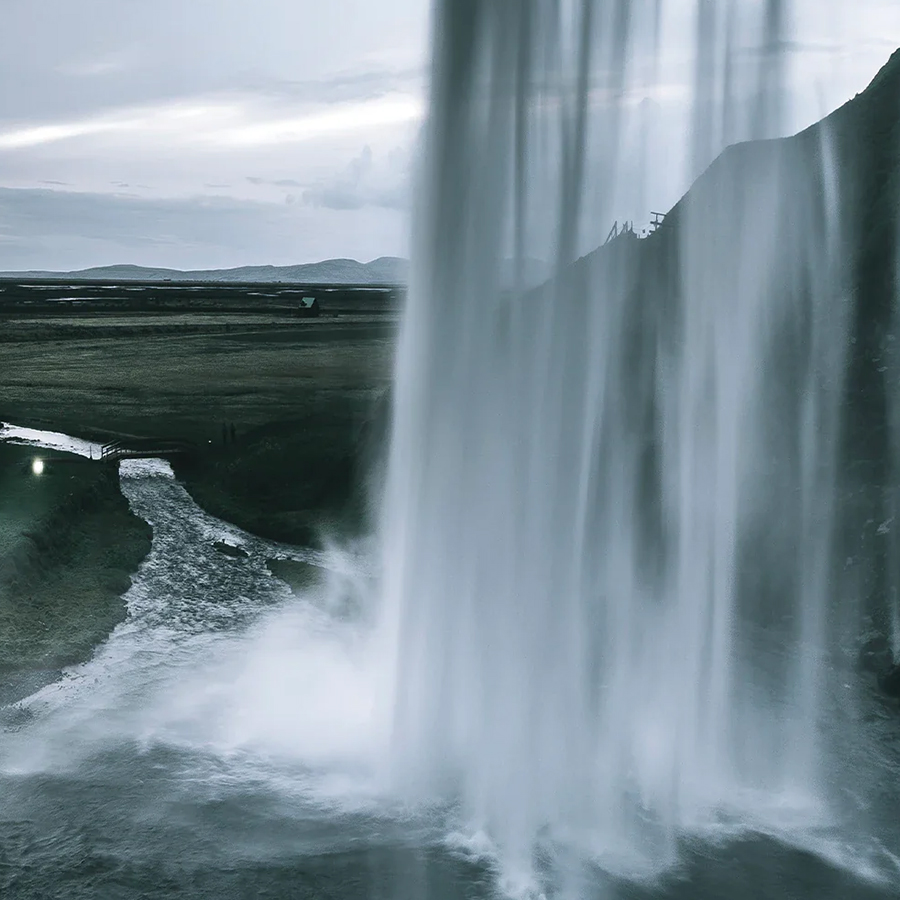 A close-up view of a waterfall cascading over a rock ledge, with mist rising and a river flowing below. The landscape features rolling hills in the background under a cloudy sky.