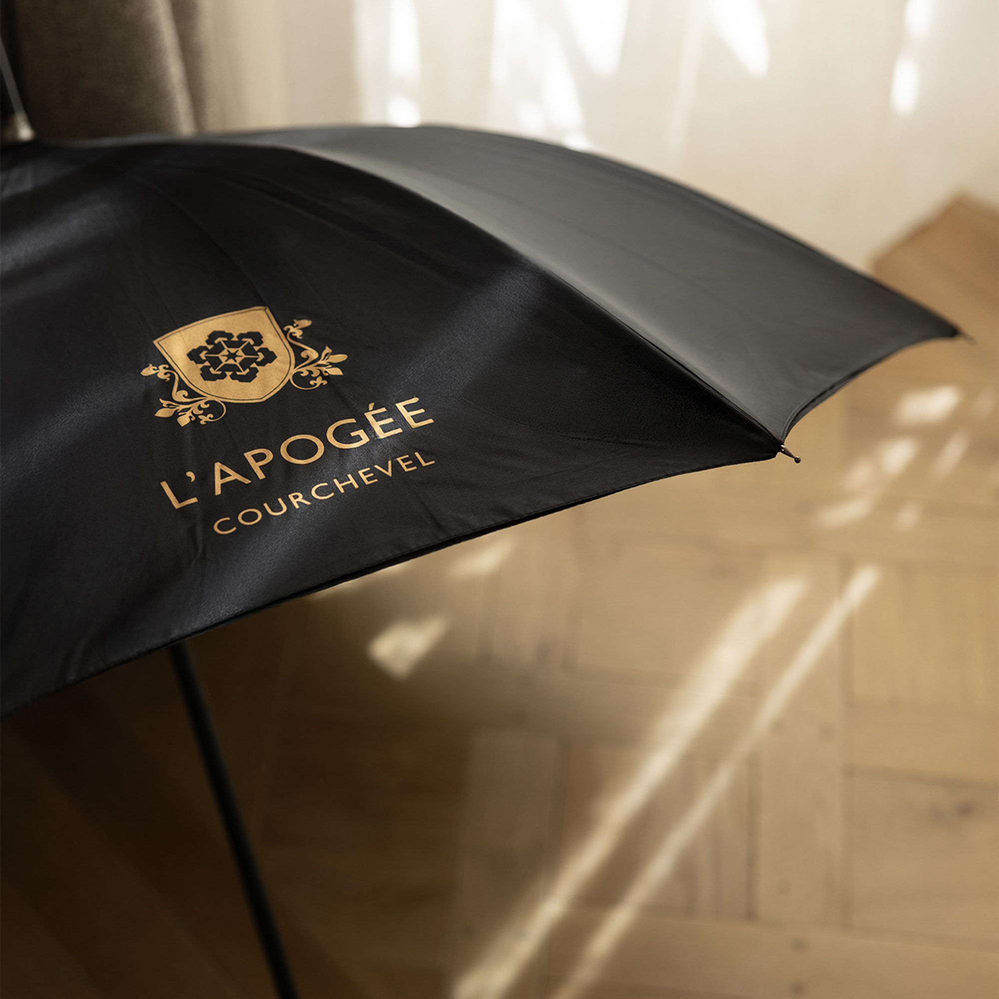 A black umbrella featuring a gold logo for "L'Apogée Courchevel" rests on a wooden floor, with sunlight casting shadows on the surface.