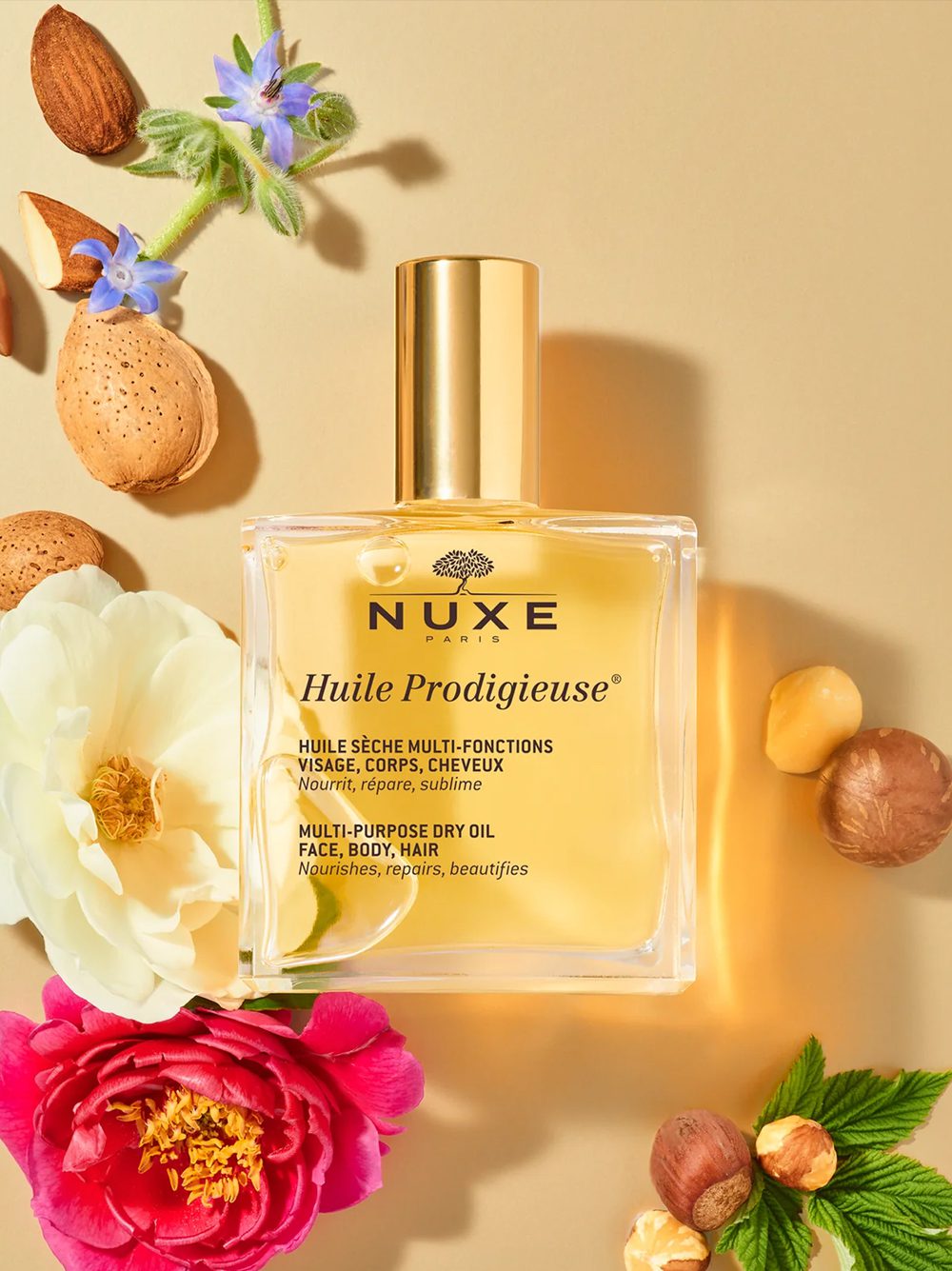 A bottle of NUXE Huile Prodigieuse multi-purpose dry oil surrounded by colorful flowers, almonds, and petals on a golden background.