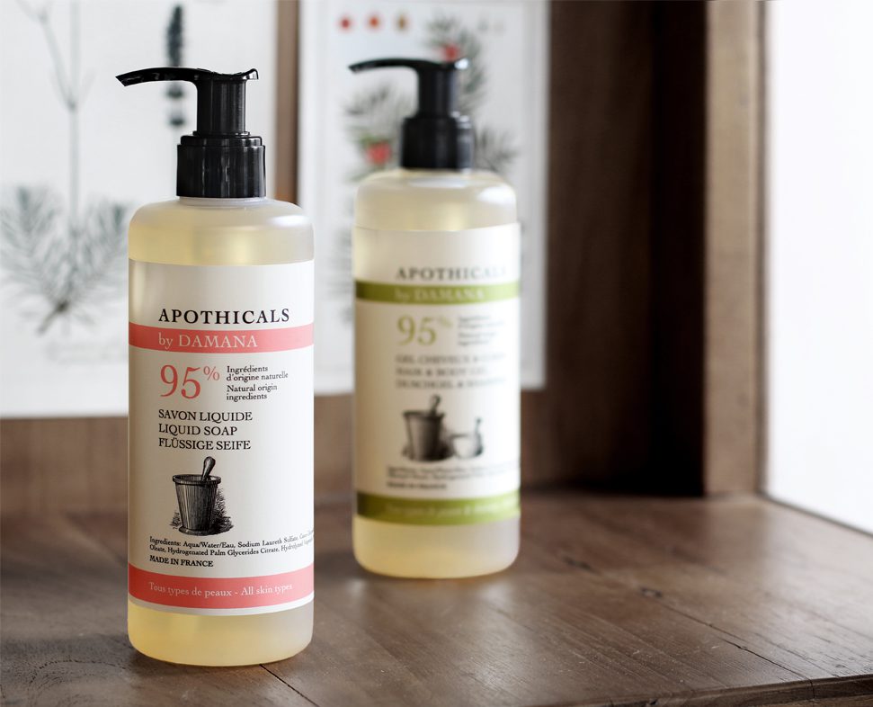 Two liquid soap bottles with pump tops, one labeled in red and the other in green, displayed on a wooden shelf against a backdrop of botanical illustrations. Both bottles feature text indicating they contain 95% natural ingredients.