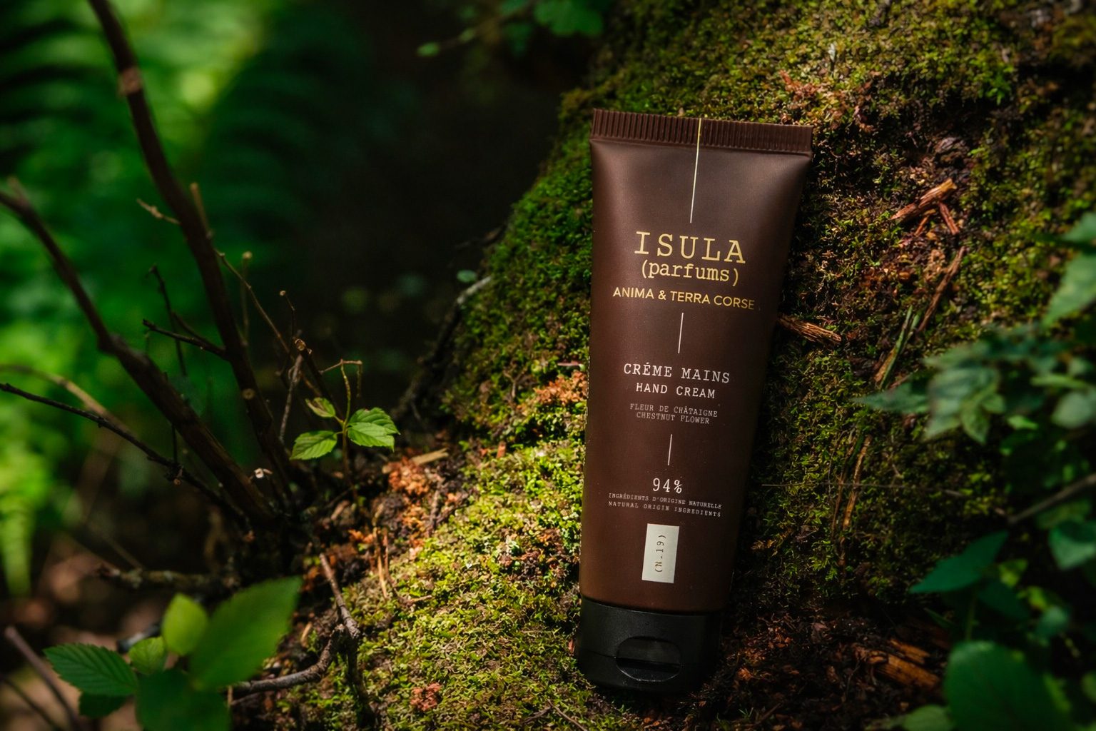 A tube of Isula hand cream rests on a moss-covered log surrounded by green foliage. The packaging is brown with gold text, indicating its natural ingredients.