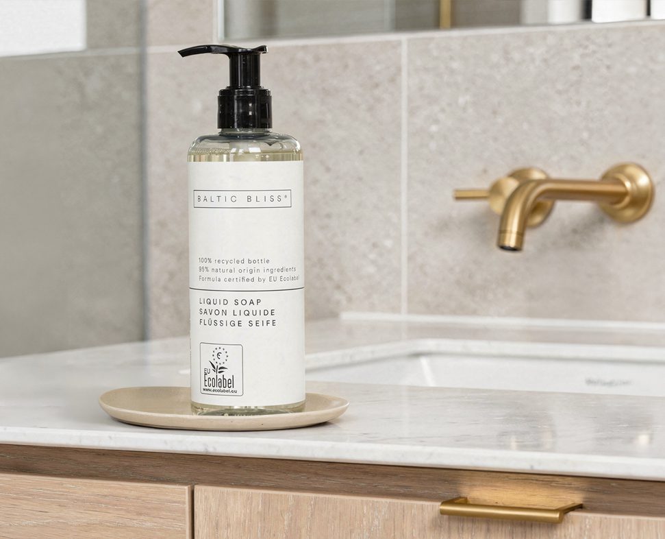 A bottle of liquid soap labeled "Baltic Bliss" sits on a small beige dish on a marble countertop, with a stylish gold faucet in the background. The bottle features a pump top and indicates it is made from 100% recycled materials.