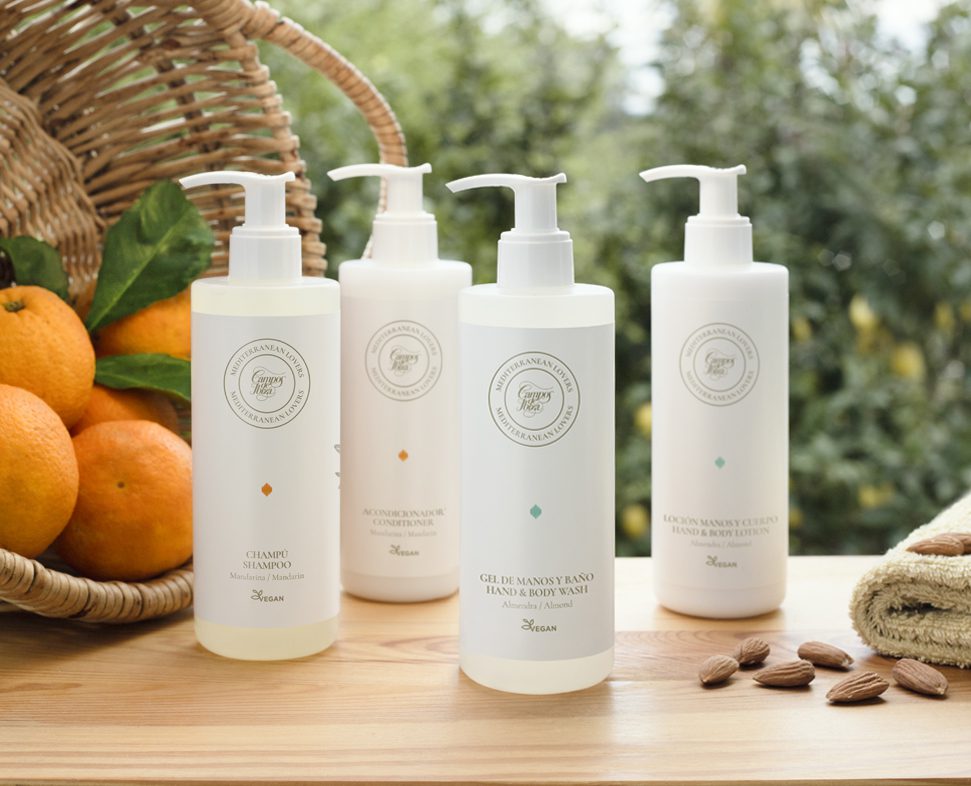 A collection of three bottles of hair and body care products displayed on a wooden surface, with oranges and almonds in the background. The bottles have a minimalist white design and feature labels in a circular format.