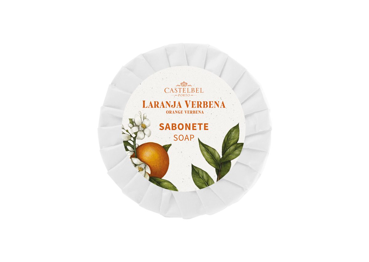 A round, white soap bar with a decorative label featuring orange and green leaves, labeled "Castelbel Laranja Verbena Soap." The design includes an illustration of an orange.