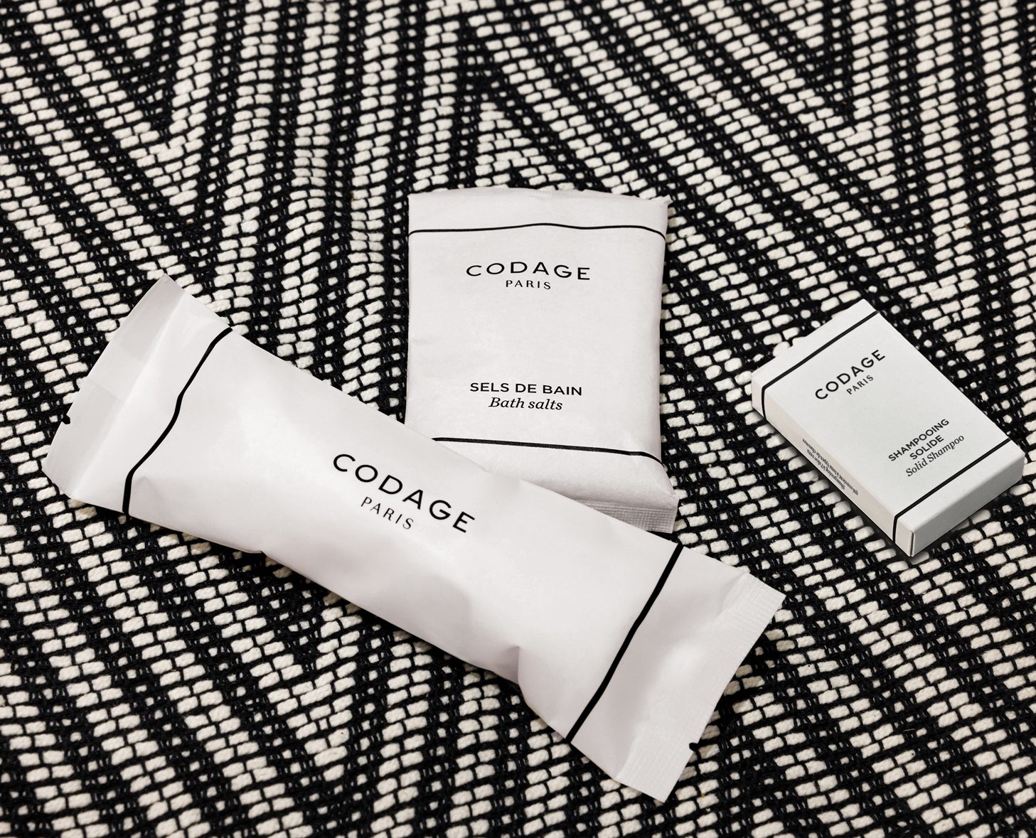 Three CODAGE Paris bath products, including a small package of bath salts and a bar of shampoo, displayed on a patterned black and white background.