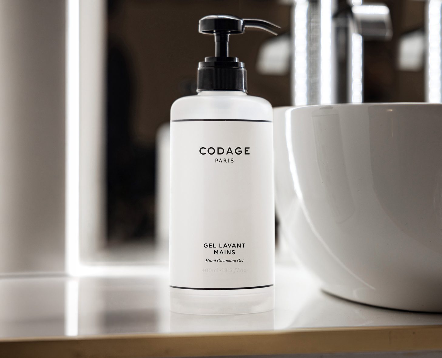 A clear bottle of CODAGE Paris hand cleansing gel with a pump dispenser, placed next to a white sink and illuminated by soft lighting.
