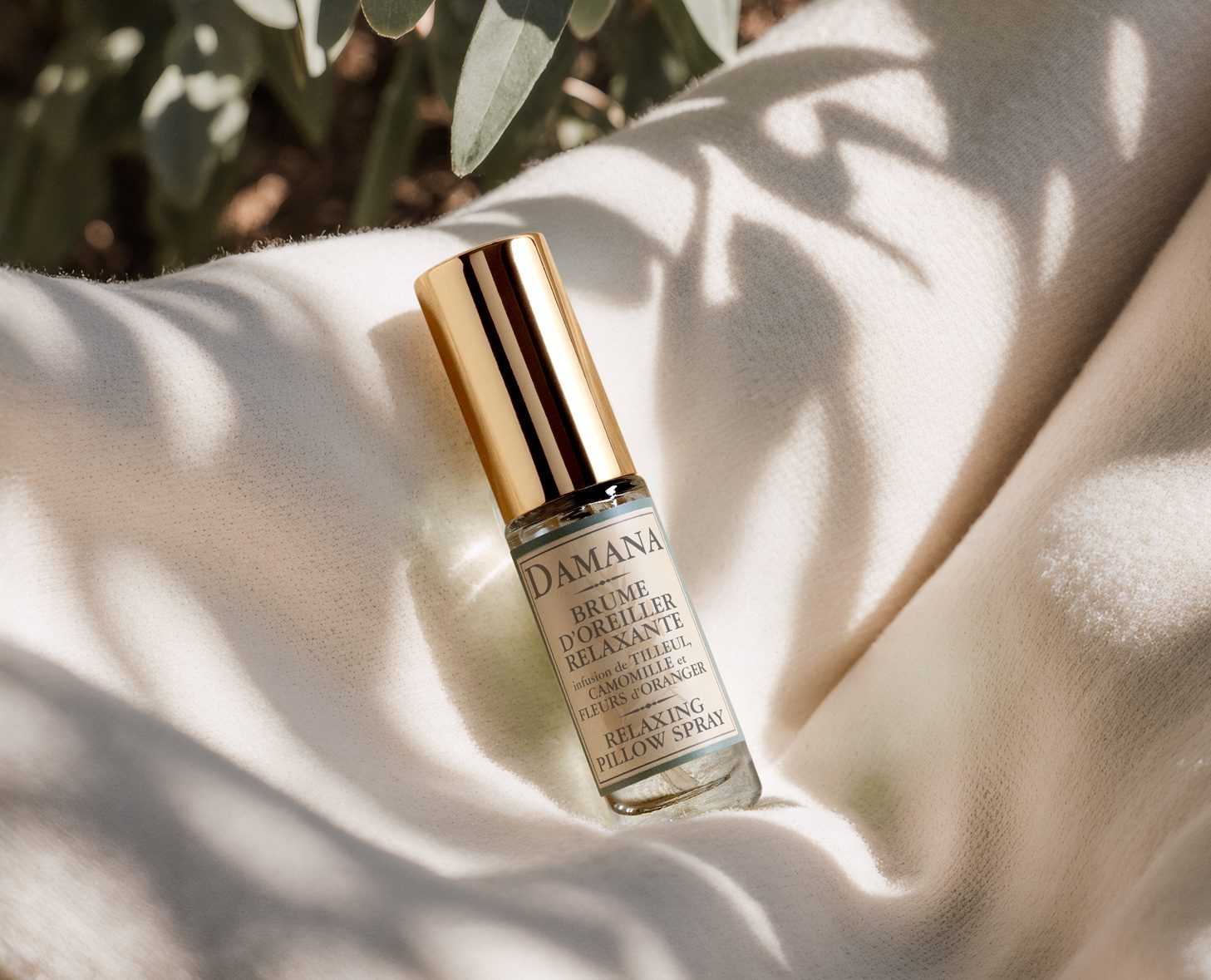 A small glass bottle of Damana Relaxing Pillow Spray with a gold cap resting on a light-colored fabric, surrounded by soft green foliage.