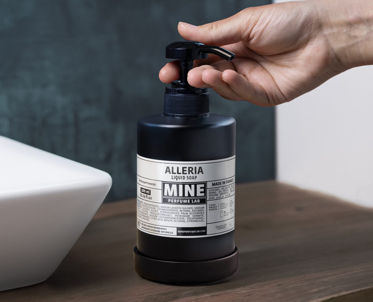 A hand is holding a pump bottle of Alleria liquid soap labeled "MINE" on a wooden surface, next to a white sink. The bottle features a modern design with a black and white label.