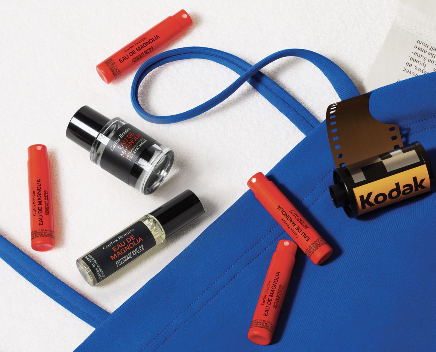 A blue tote bag surrounded by various cosmetics, including red lip products and a bottle of perfume, along with a roll of Kodak film.