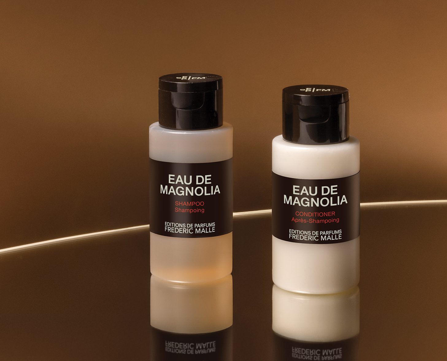 A bottle of Eau de Magnolia shampoo and a bottle of Eau de Magnolia conditioner, both elegantly labeled, placed on a reflective surface against a brown background.