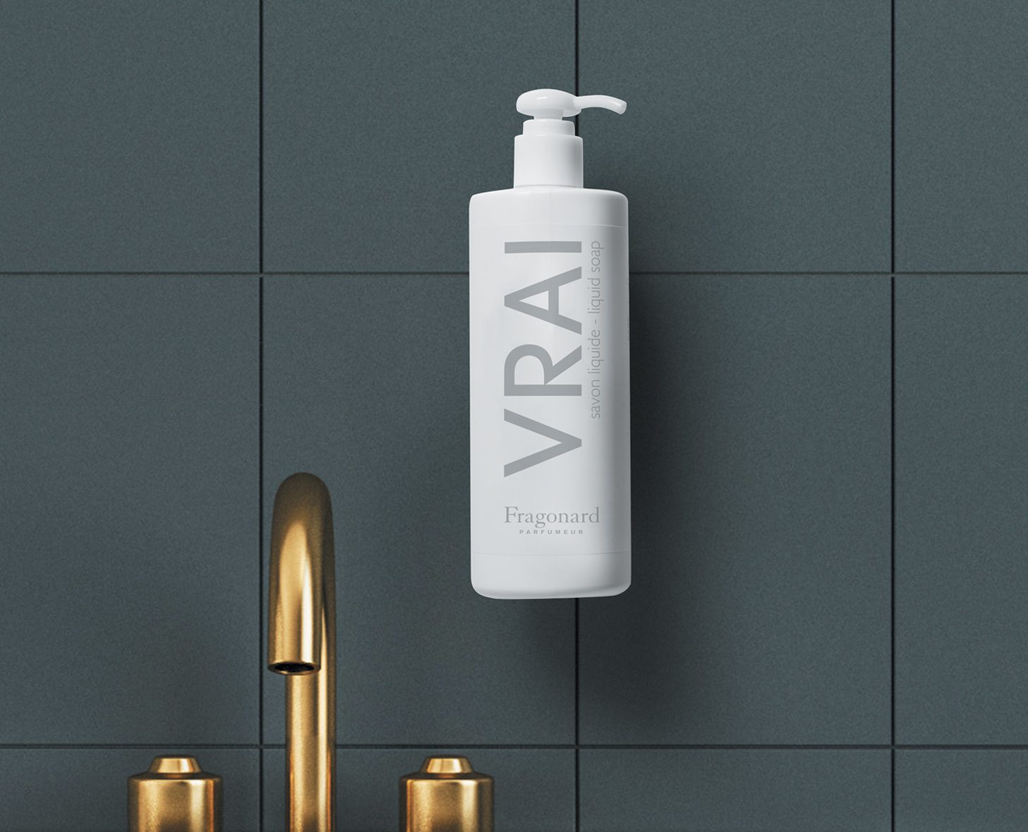 A white pump bottle labeled "VRAI" with a minimalist design, mounted on a dark tiled wall beside a gold faucet.