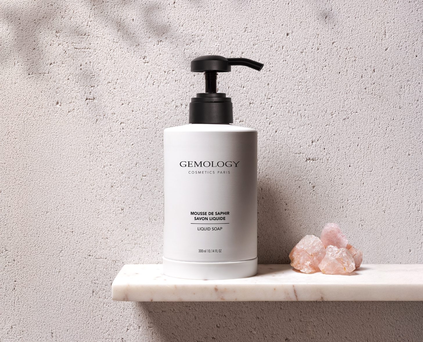 A white bottle of Gemology liquid soap with a black pump sits on a marble shelf, accompanied by pink crystals against a textured gray wall.
