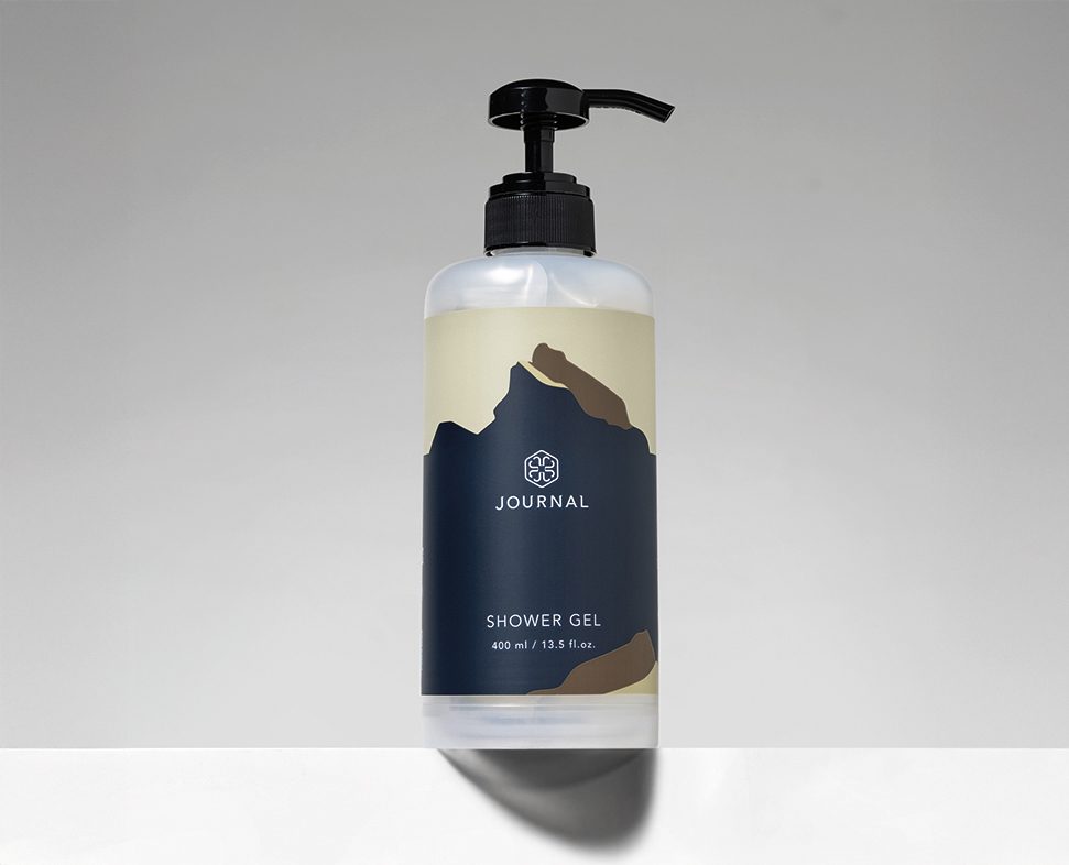 A transparent bottle of shower gel with a pump dispenser, featuring a minimalist label design that includes mountain graphics and the text "JOURNAL SHOWER GEL."