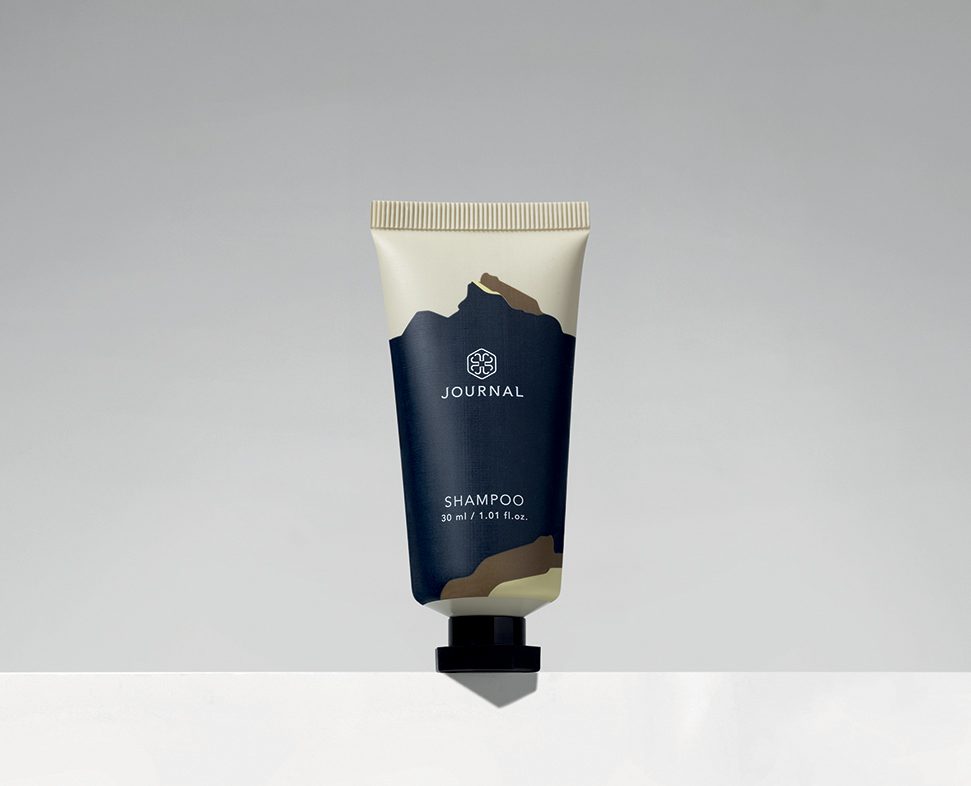 A small tube of shampoo labeled "JOURNAL" with a minimalist design featuring a dark blue and cream color scheme, set against a simple gray background.
