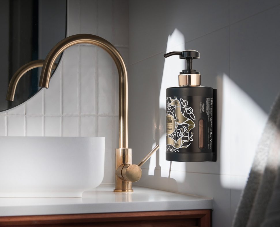 A black liquid soap dispenser with a pump is mounted on a bathroom wall beside a gold faucet and a white bowl sink, illuminated by natural light.