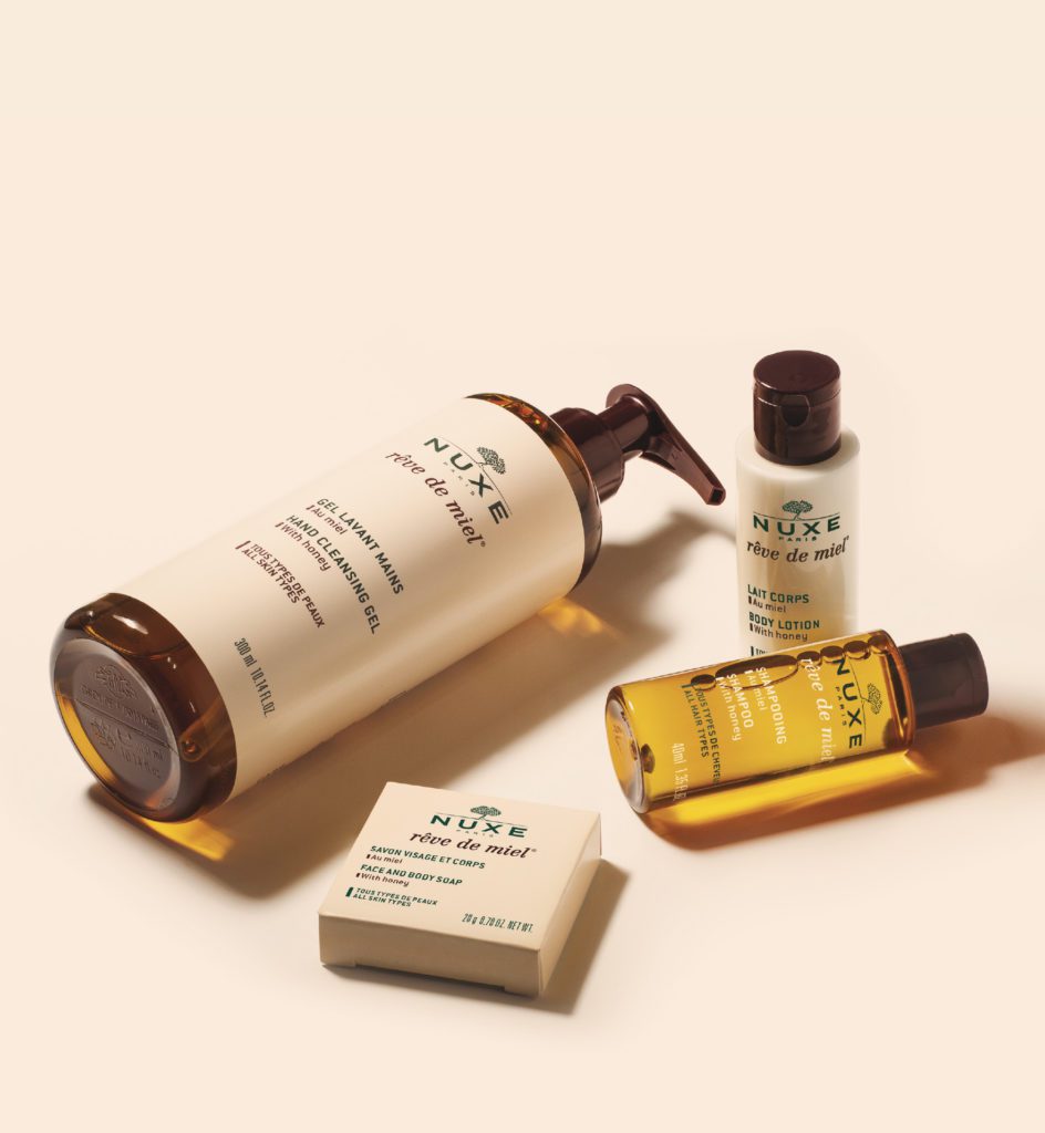 A collection of NUXE skincare products, including a large cleansing gel bottle, two smaller lotion containers, and a bar of soap, arranged on a soft pastel background.