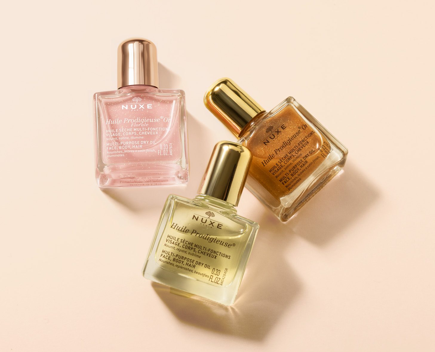 Three bottles of NUXE Huile Prodigieuse oil in pink, yellow, and gold hues against a soft beige background.