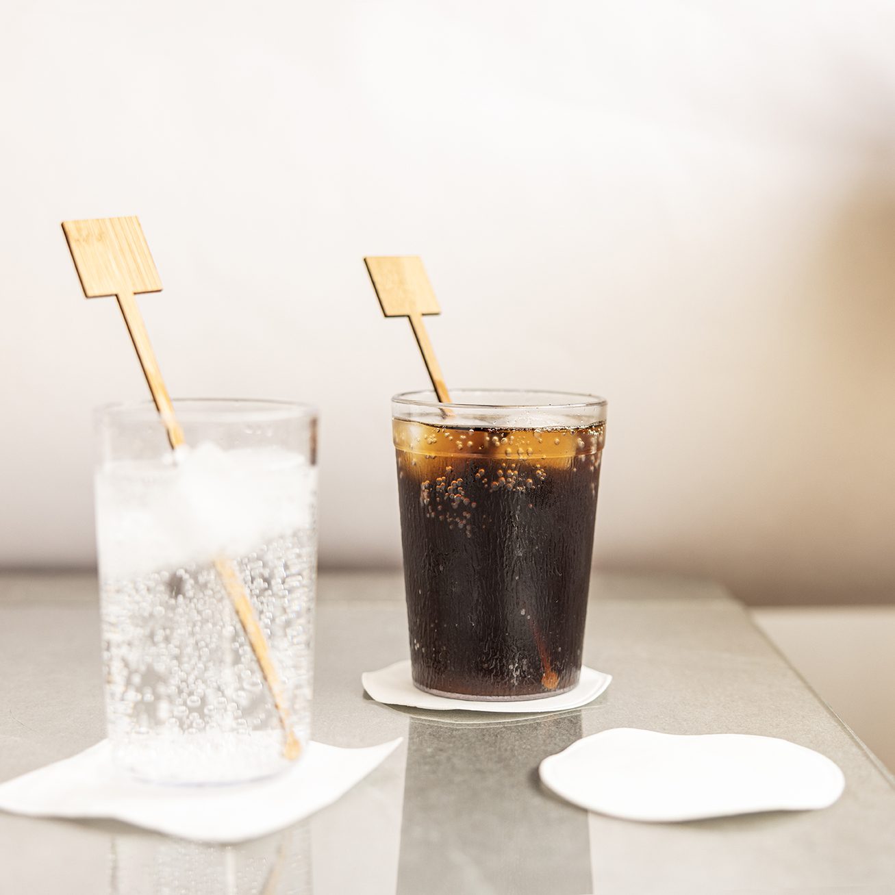 A clear glass of sparkling water with ice and a dark soda in a separate glass, both on coasters with wooden stir sticks. The background is softly blurred.