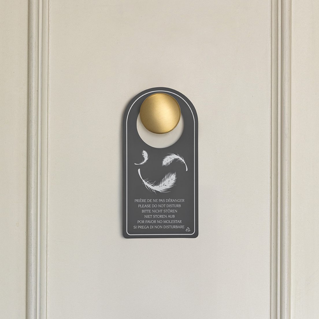A door sign featuring a gold circular knob and a gray background with text in multiple languages stating "Please Do Not Disturb" and "Do Not Disturb." The design includes faint feathers or decorative elements.