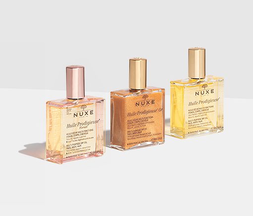 Three bottles of NUXE Huile Prodigieuse oils, featuring pink, gold, and yellow liquid, displayed on a light background. Each bottle has a label and a gold cap.