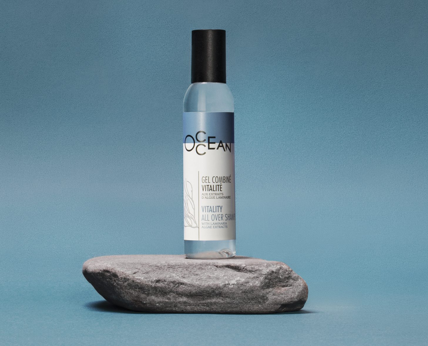 A bottle of "OCEAN" vitality gel stands upright on a gray rock against a blue background. The label features a minimalist design with botanical illustrations.