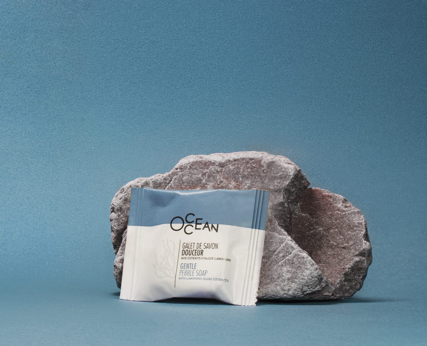 A bar of gentle pebble soap is placed next to a textured stone against a solid blue background, showcasing its packaging with the label "OCEAN."