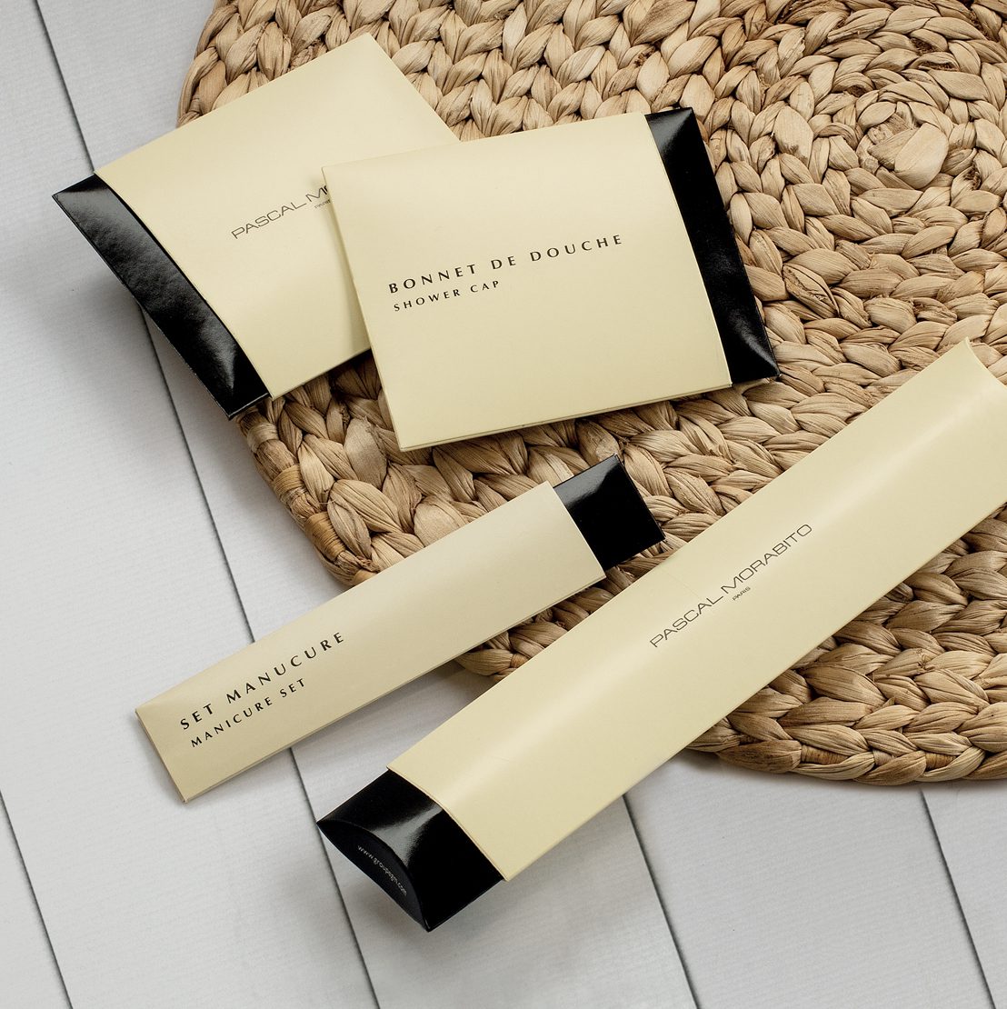 A flat lay of a shower cap packaging, a manicure set, and a body wash package arranged on a woven mat. The items feature a minimalist design with elegant typography.