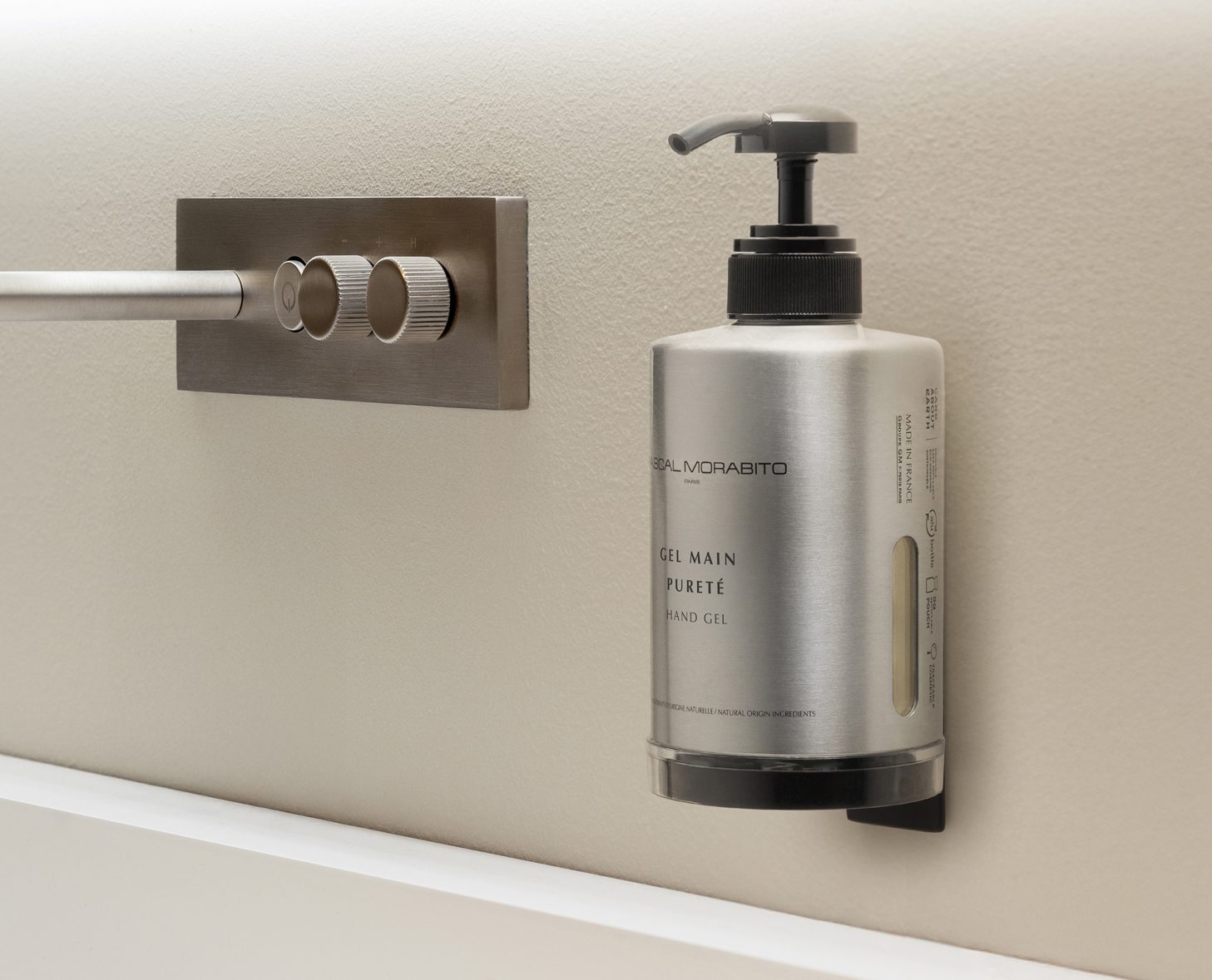 A stainless steel dispenser of hand gel mounted on a wall next to a control knob for water temperature. The dispenser features a pump top and clearly labeled contents.