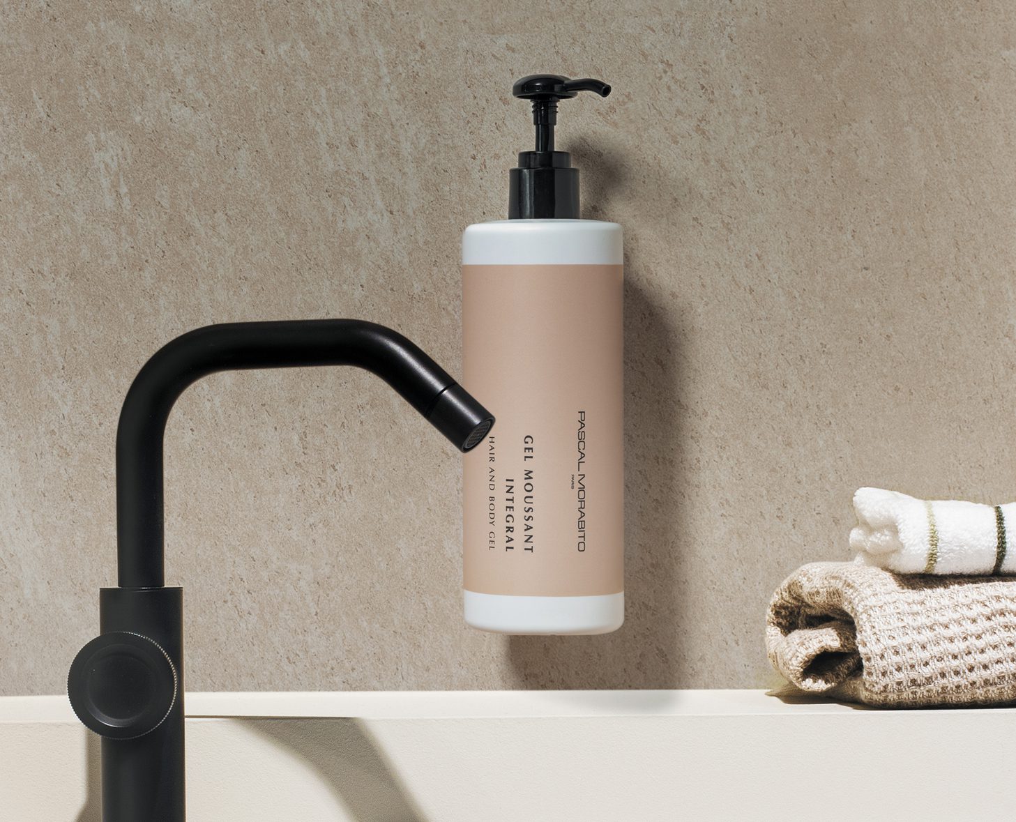 A pump bottle of cleansing gel is mounted on a light stone wall next to a black faucet and a folded cream-colored towel.