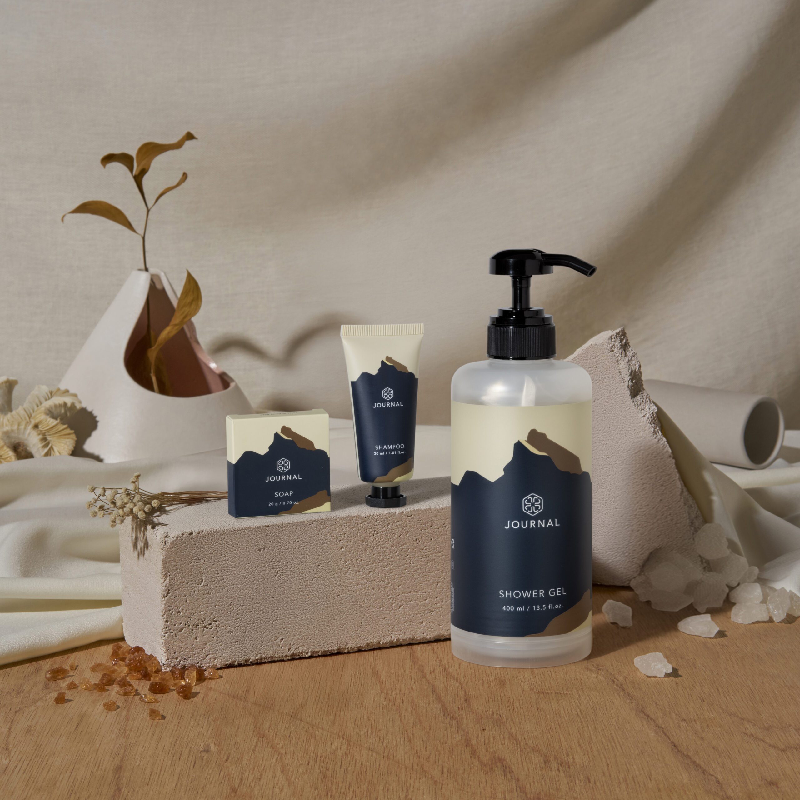 A collection of minimalist beauty and grooming products displayed on a textured surface, including a shower gel bottle, a shampoo tube, and a bar soap, with a neutral background featuring abstract shapes and dried plants.